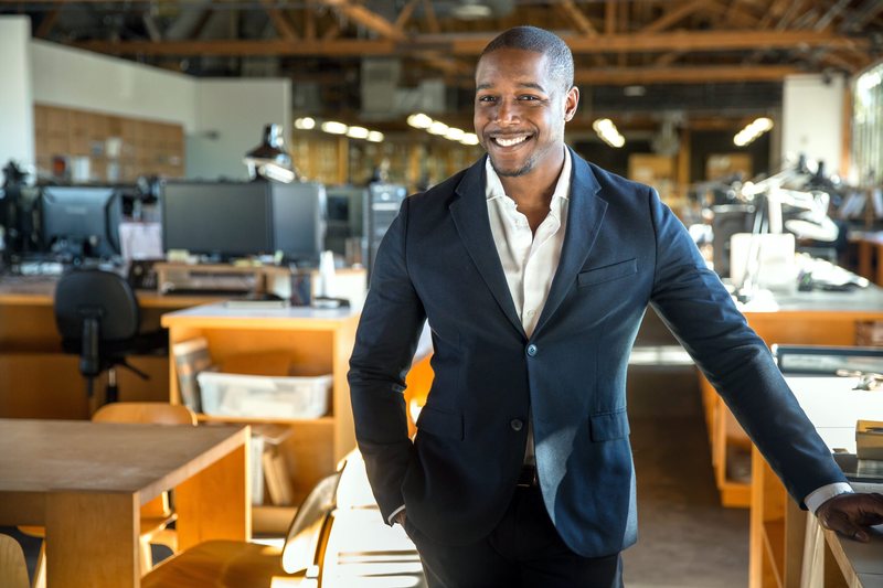 A businessman in a dark suit smiling in a bright open-plan office.