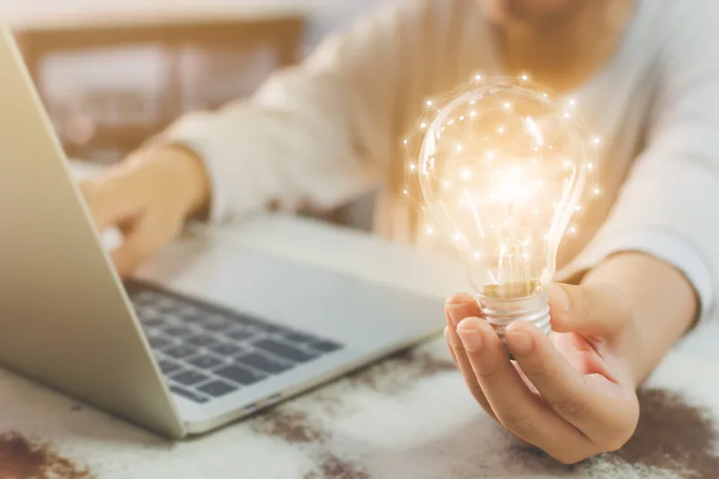 A person at a laptop holds a glowing light bulb in one open palm, with a network of dots and lines overlaid on the bulb.
