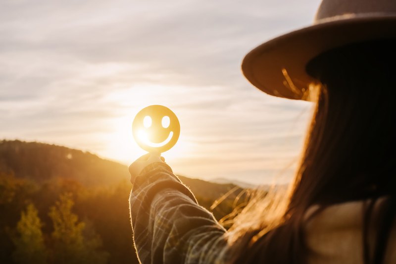 A person in a wide-brimmed hat holds up a smiley face cutout against the sun setting over wooded hills.