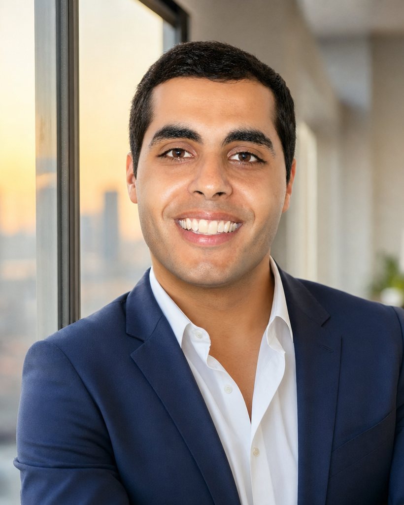 Mark Elbadramany, arms folded, in a navy suit jacket and open white shirt, photographed beside a floor-to-ceiling window with a city skyline at sunset behind him.