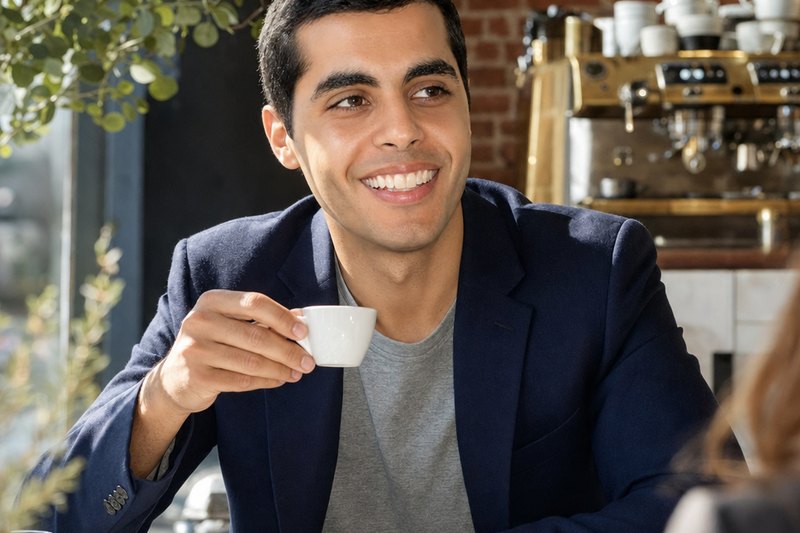 A smiling man in a navy blazer and grey T-shirt holds a small white espresso cup while seated at a marble table in a cafe with an espresso machine and brick wall behind him.